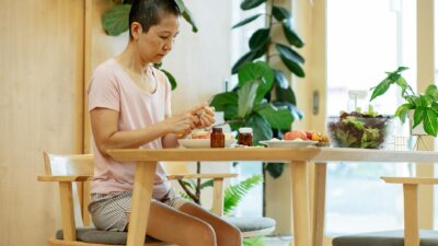 Serious adult ethnic woman with short dark hair in comfy clothes sitting at wooden table with bowl of salad and medicines while peeling healthy fruits at home
