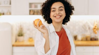 A joyful woman with curly hair holding an orange in her kitchen, promoting a healthy lifestyle.
