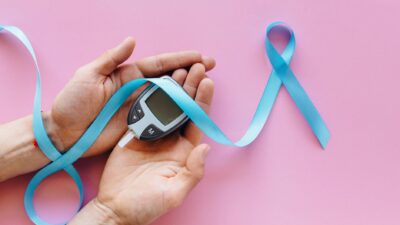 A close-up image of hands holding a glucometer with a blue awareness ribbon on a pink surface, symbolizing diabetes awareness.