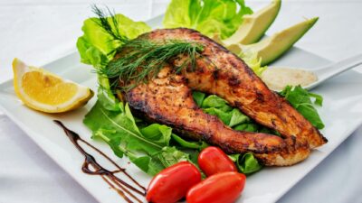 Delicious grilled salmon steak served with avocado, lettuce, cherry tomatoes, and lemon slice on a white plate.
