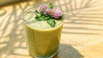 Close-up of a nutritious green smoothie topped with fresh purple clover flowers in sunlight.