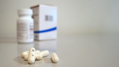 Close-up image of white capsules with medicine bottle and box in the background.