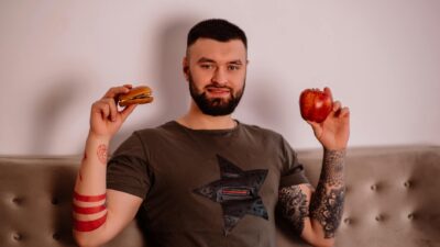 Bearded man pondering diet choice with apple and burger. Indoors with tattoos visible.