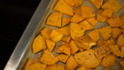 From above of appetizing sliced fried potatoes with spices scattered on oven pan in kitchen
