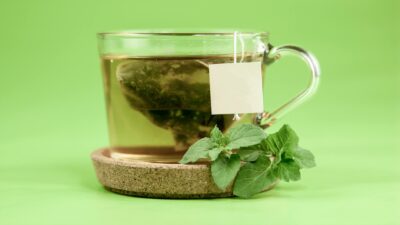 Green tea bag in glass cup with hot water on cup holder with mint leaves placed on green background