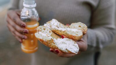 Close-up of hands holding a bagel with cream cheese and a bottle of orange juice outdoors.