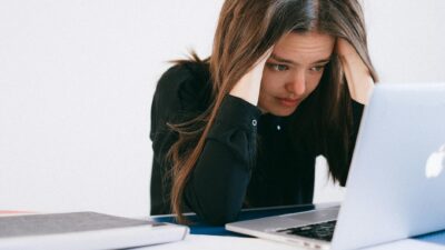 A stressed woman at a desk, looking at a laptop with a worried expression.