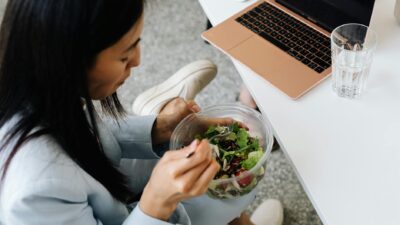 Woman eating a fresh salad at her desk with a laptop nearby, promoting healthy eating at work.